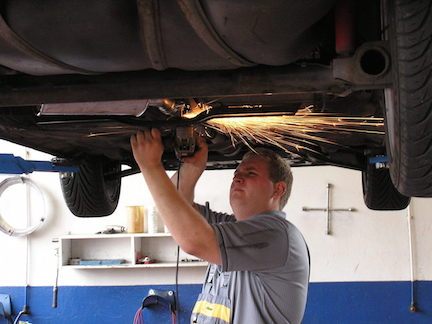 An auto mechanic repairing a muffler on a car, using a cutting tool which is creating sparks.