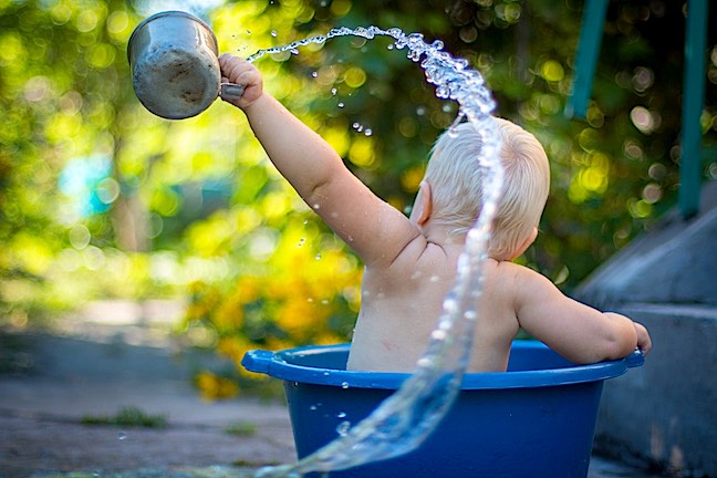 A baby playing in a small pool splashing water from a cup.