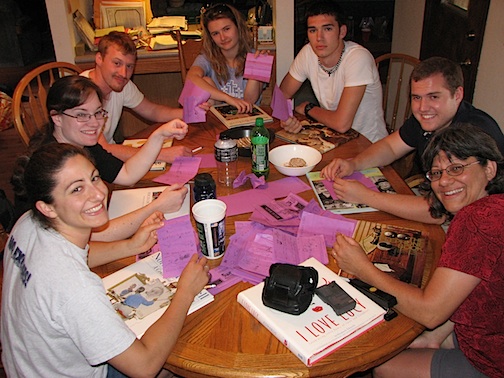 Family Game Night Ideas: 46 Activities and Games That Are Fun & Cheap 6 A group of people sitting around a table on game night playing telephone pictionary.