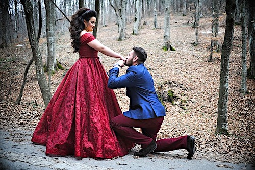 Man in a blue jacket on his knee proposing to a woman in a scarlet gown.